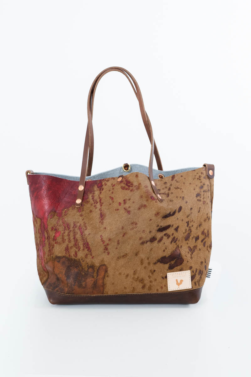 A tote bag featuring a mottled brown and beige cowhide pattern, a red painted section on the left, a light blue interior lining, and dark brown leather handles and trim, shown on a white background. Handcrafted by Meanwhile Back on the Farm in Lynchburg, Va.