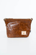 A brown distressed leather crossbody bag with a small beige patch featuring a heart logo on the front, set against a plain white background. Handcrafted by Meanwhile Back on the Farm in Lynchburg, Va.