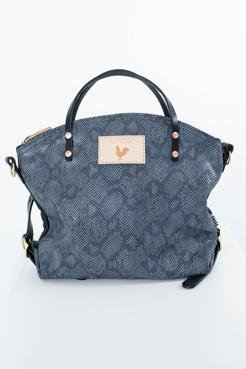 A blue-gray snakeskin-patterned handbag with black leather handles and a beige rectangular patch featuring a small rooster logo, photographed against a plain white background. Handcrafted by Meanwhile Back on the Farm in Lynchburg, Va.