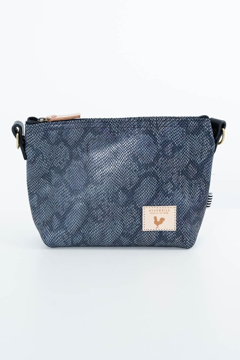 A small crossbody bag with a dark blue-gray snakeskin pattern, featuring a tan leather brand patch on the front and a black strap. Handcrafted by Meanwhile Back on the Farm in Lynchburg, Va.