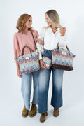 Two women standing side by side against a white studio background, each holding a matching patterned tote bag and a matching zip pouch. The women are wearing different outfits, including a pink sweater, a white blouse, and blue jeans, and both bags feature a multicolor paisley-like pattern with brown handles and trims. Handcrafted by Meanwhile Back on the Farm in Lynchburg, Va.