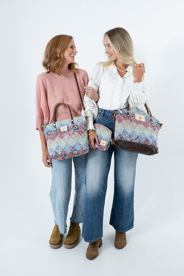 Two women standing against a white studio background, each holding a matching quilt-patterned tote bag and clutch set. The women are dressed in casual outfits, one in a pink sweater and blue jeans, and the other in a white blouse and blue jeans. Handcrafted by Meanwhile Back on the Farm in Lynchburg, VA.