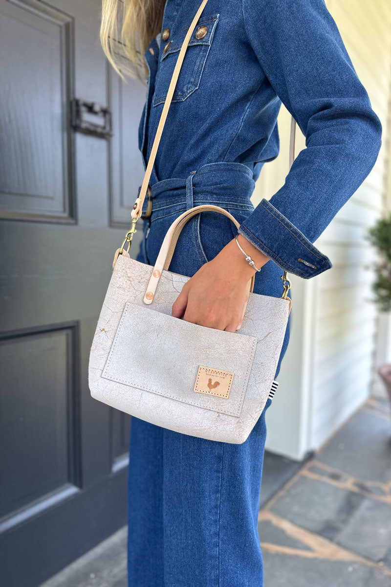 Woman holding the birch white shoulder strap bag with the meanwhile logo and a white body strap attached to the bag.