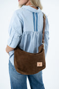 A woman wearing a light blue blouse and jeans carries a brown suede shoulder bag with a tan leather patch on a white background. Handcrafted by Meanwhile Back on the Farm in Lynchburg, VA.