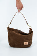 A brown suede shoulder bag with a matching leather strap and gold-tone hardware, held by a hand against a plain white background. Handcrafted by Meanwhile Back on the Farm in Lynchburg, VA.