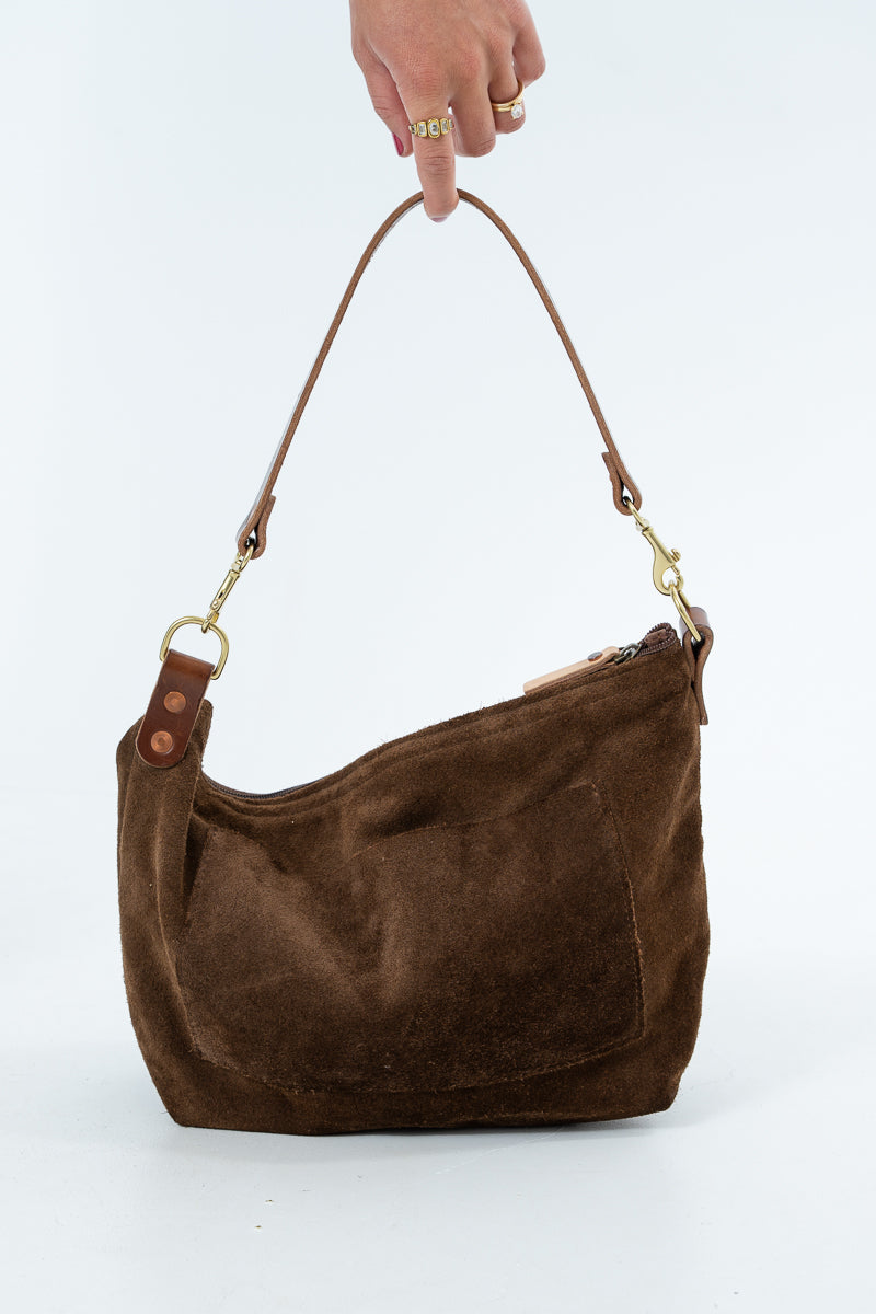 A brown suede shoulder bag with a matching leather strap and gold-tone hardware, held by a hand against a plain white background. Handcrafted by Meanwhile Back on the Farm in Lynchburg, VA.