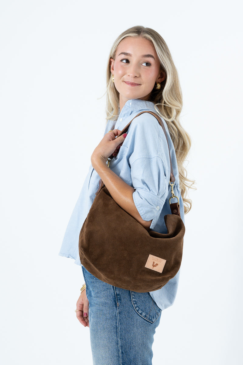 A woman with blonde hair wearing a light blue shirt and jeans, carrying a brown suede crescent-shaped shoulder bag with a small tan leather logo patch. Handcrafted by Meanwhile Back on the Farm in Lynchburg, VA.