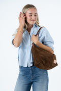 A woman wearing a light blue button-up shirt and blue jeans carries a brown suede shoulder bag with a leather strap against a plain white background. Handcrafted by Meanwhile Back on the Farm in Lynchburg, VA.