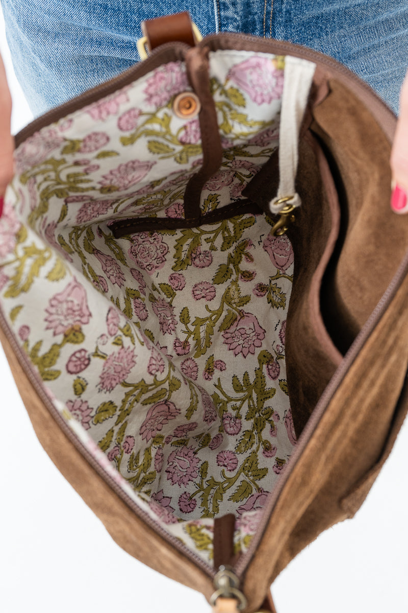 Close-up of an open brown suede or canvas bag showing a floral-patterned interior lining with pink flowers and green stems on a light background. A hand with pink nail polish is visible holding the bag, and part of blue jeans is visible at the top. Handcrafted by Meanwhile Back on the Farm in Lynchburg, VA.
