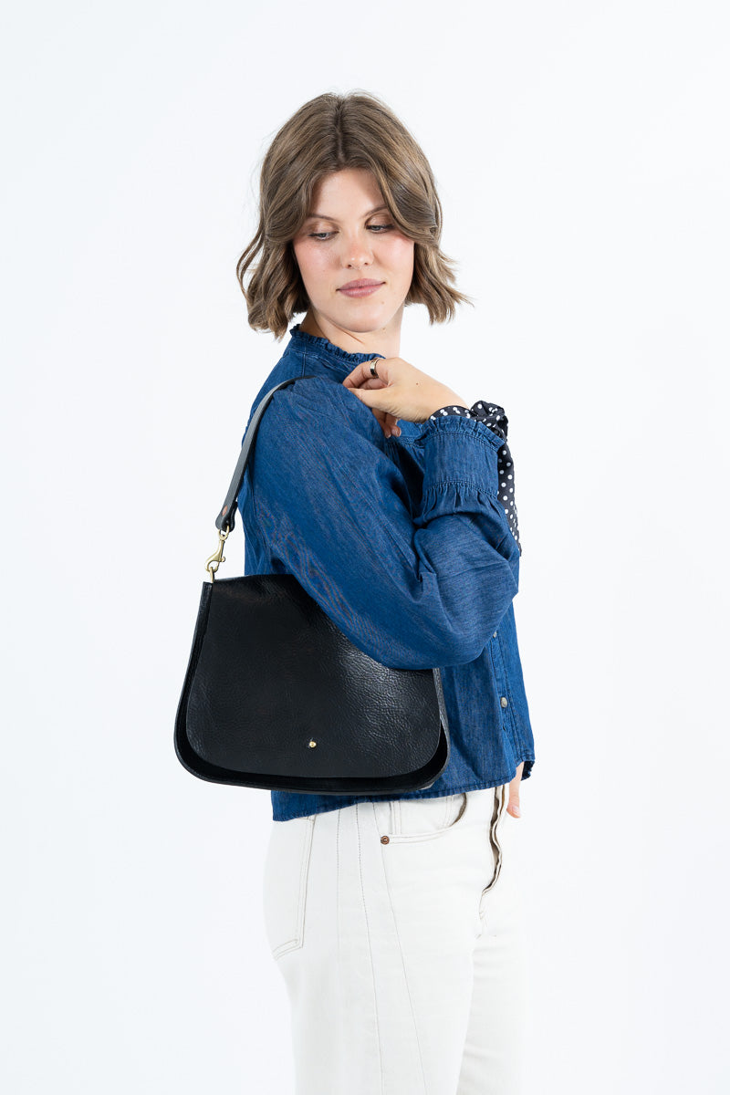 A woman wearing a blue denim jacket and white pants carries a black leather shoulder bag with gold hardware against a plain white background. Handcrafted in Lynchburg, VA.