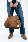 A person wearing blue jeans and brown suede boots holds a large cowhide-textured shoulder bag with brown leather handles and trim against a plain white background. Handcrafted by Meanwhile Back on the Farm in Lynchburg, Va.