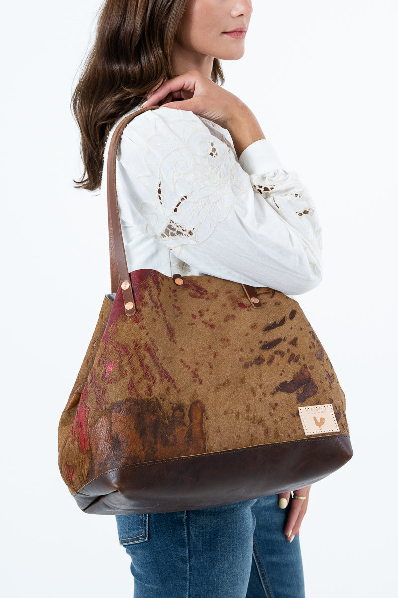 A woman wearing a white embroidered blouse and blue jeans carries a large brown leather shoulder bag with a distressed, mottled tan and reddish-brown textured body and dark brown leather trim. Handcrafted by Meanwhile Back on the Farm in Lynchburg, Va.