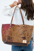 A woman holding a large brown and red mottled leather tote bag with dark brown handles and a small tan brand patch on the bottom right, against a plain white background. Handcrafted by Meanwhile Back on the Farm in Lynchburg, Va.