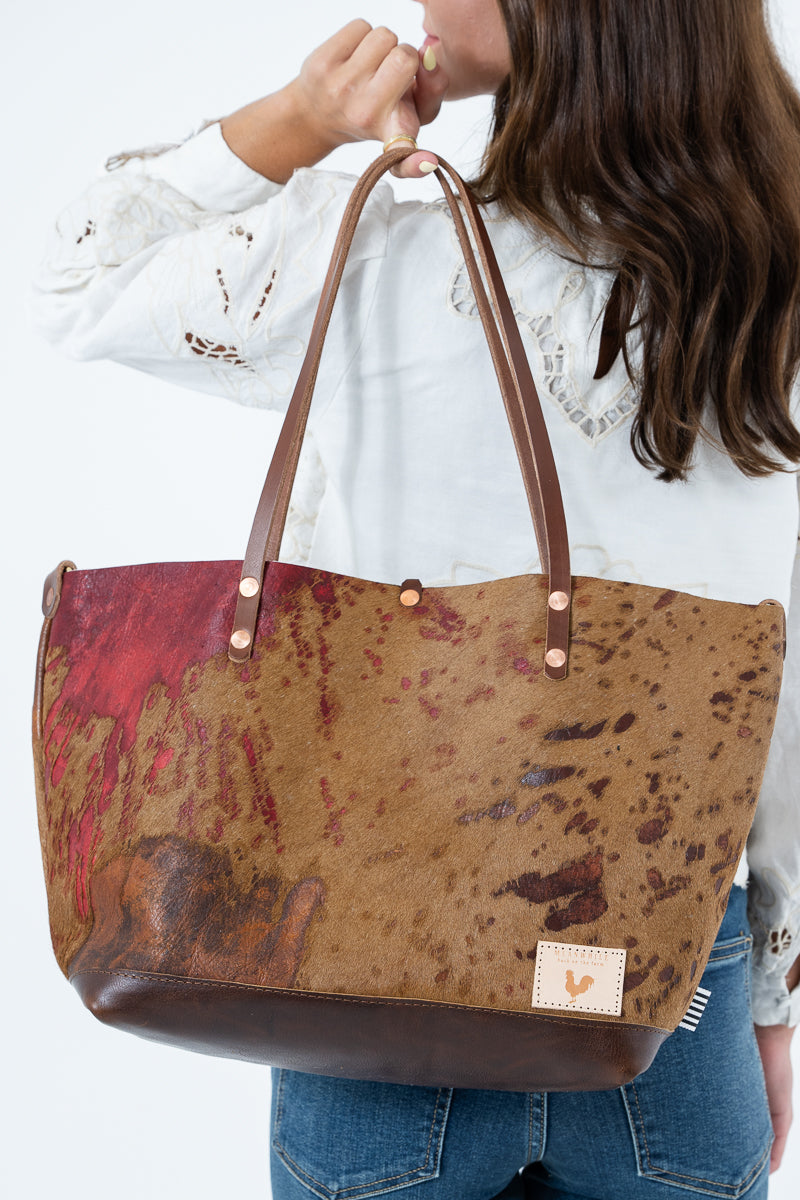 A woman holding a large brown and red mottled leather tote bag with dark brown handles and a small tan brand patch on the bottom right, against a plain white background. Handcrafted by Meanwhile Back on the Farm in Lynchburg, Va.