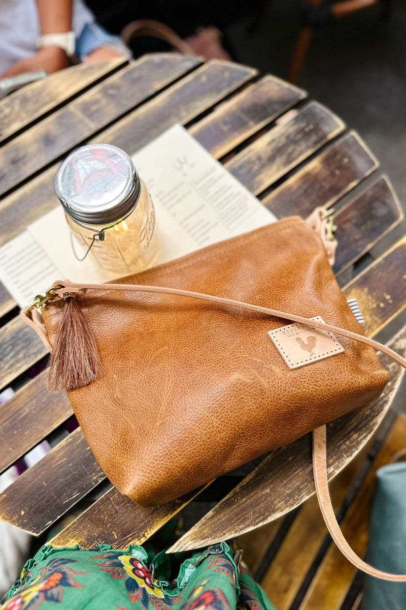 A camel brown bag with meanwhile logo, cross body strap, and a brown tassel zipper lying on a wood table. 