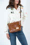 A woman wearing a white embroidered button-up shirt and blue jeans carries a brown faux-leather crossbody bag with a tan rooster logo patch and a brown strap. Handcrafted by Meanwhile Back on the Farm in Lynchburg, Va.