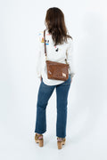A woman viewed from behind wearing a white embroidered long-sleeve top, blue jeans, and brown platform sandals, while carrying a brown leather messenger bag over her shoulder against a white studio background. Handcrafted by Meanwhile Back on the Farm in Lynchburg, Va.