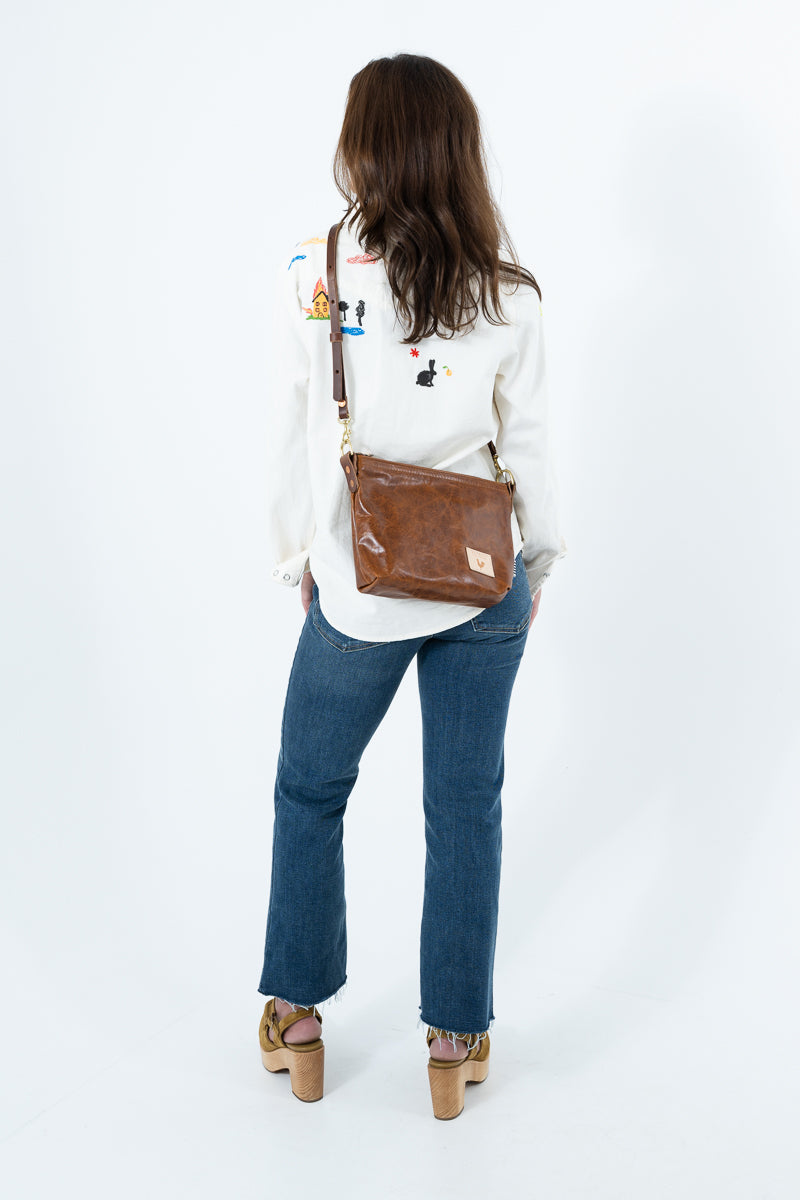 A woman viewed from behind wearing a white embroidered long-sleeve top, blue jeans, and brown platform sandals, while carrying a brown leather messenger bag over her shoulder against a white studio background. Handcrafted by Meanwhile Back on the Farm in Lynchburg, Va.
