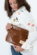 A woman holding a brown leather clutch bag with a small tan patch featuring a rooster logo. She is wearing a white embroidered button-up shirt and blue jeans against a plain white background. Handcrafted by Meanwhile Back on the Farm in Lynchburg, Va.