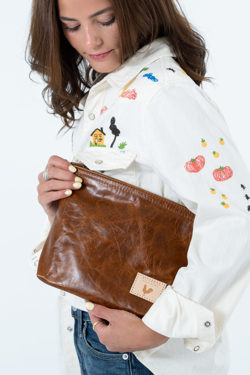 A woman holding a brown leather clutch bag with a small tan patch featuring a rooster logo. She is wearing a white embroidered button-up shirt and blue jeans against a plain white background. Handcrafted by Meanwhile Back on the Farm in Lynchburg, Va.