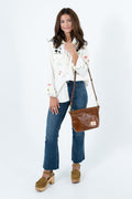 A woman modeling a white embroidered long-sleeve blouse, blue jeans, and brown platform clogs, carrying a brown leather shoulder bag against a white studio background. Handcrafted by Meanwhile Back on the Farm in Lynchburg, Va.