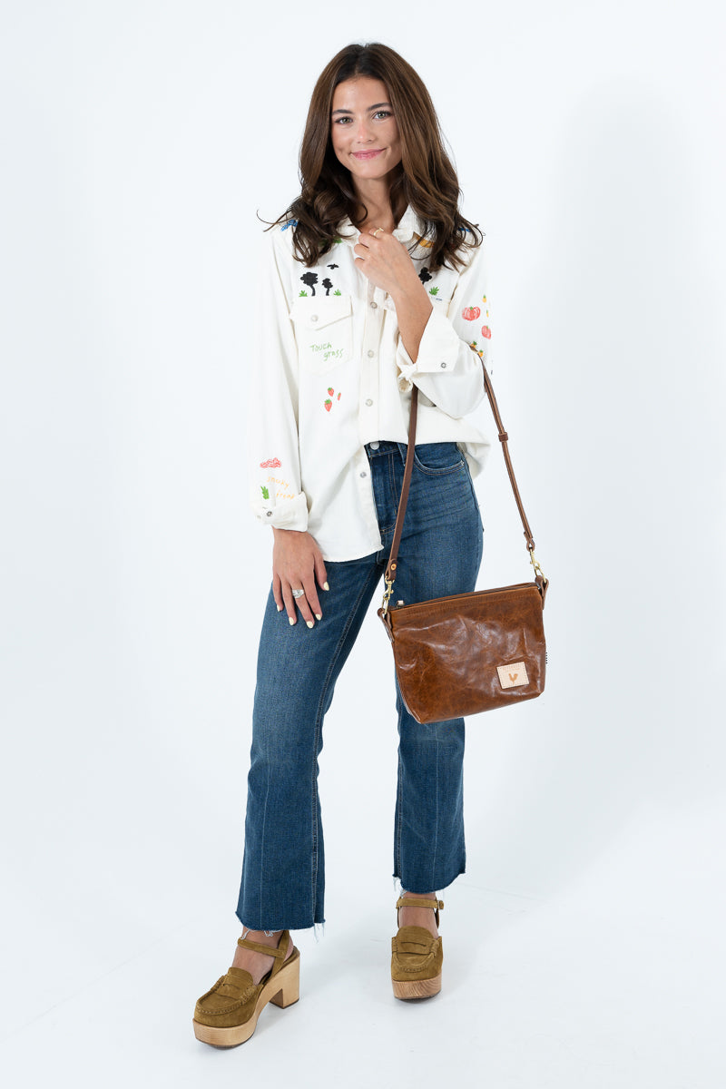 A woman modeling a white embroidered long-sleeve blouse, blue jeans, and brown platform clogs, carrying a brown leather shoulder bag against a white studio background. Handcrafted by Meanwhile Back on the Farm in Lynchburg, Va.