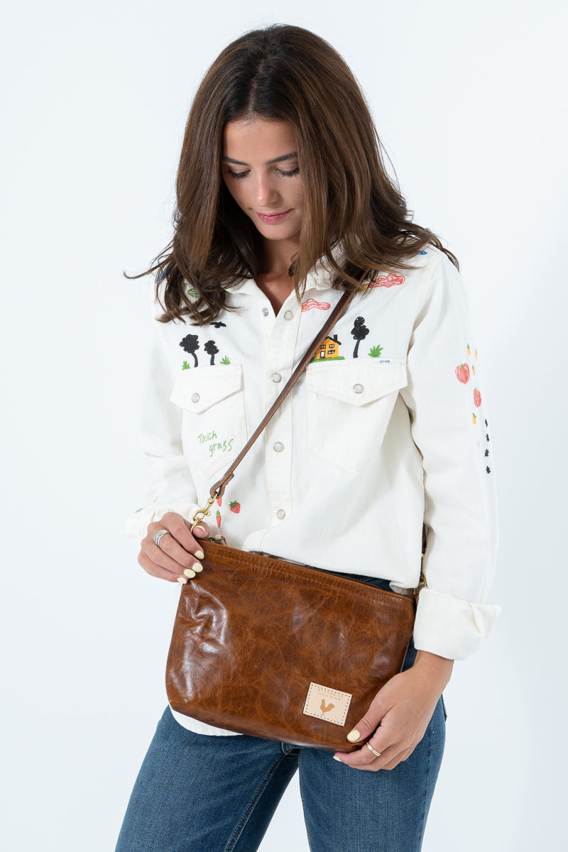 A woman wearing a white embroidered button-up shirt and blue jeans carries a brown leather crossbody bag with a matching strap. Handcrafted by Meanwhile Back on the Farm in Lynchburg, Va.