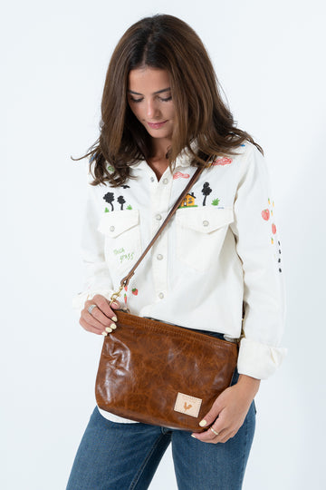 A woman wearing a white embroidered button-up shirt and blue jeans carries a brown leather crossbody bag with a matching strap. Handcrafted by Meanwhile Back on the Farm in Lynchburg, Va.