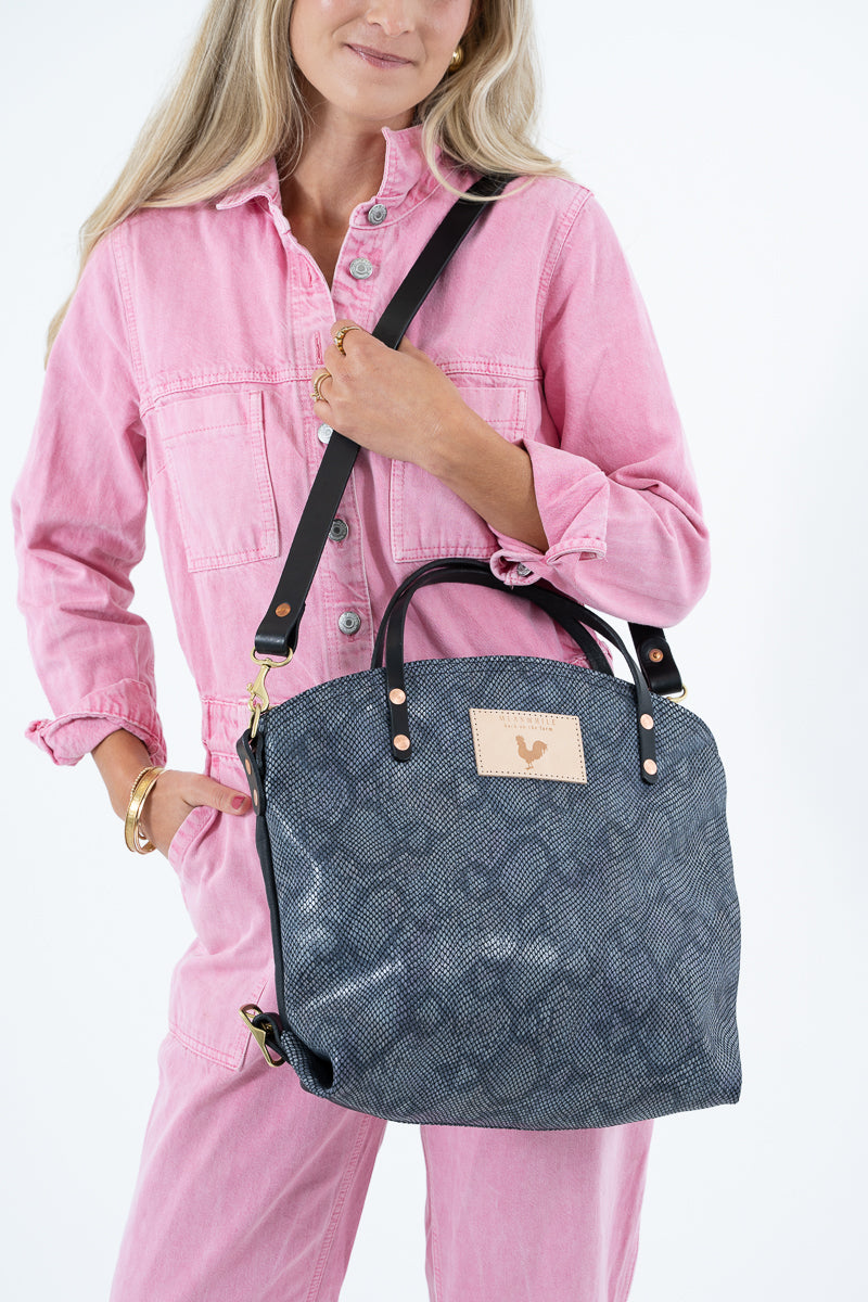 A woman wearing a pink jumpsuit carries a dark gray snakeskin leather backpack with black straps and gold-tone hardware against a plain white background. Handcrafted by Meanwhile Back on the Farm in Lynchburg, VA.