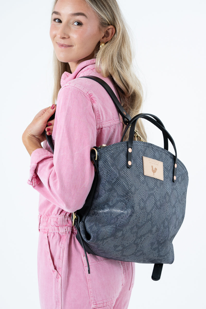 A woman wearing a pink jumpsuit carries a dark gray snakeskin leather backpack with black straps and gold-tone hardware against a plain white background. Handcrafted by Meanwhile Back on the Farm in Lynchburg, VA.
