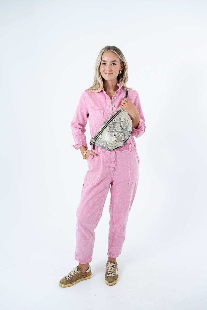 A woman wearing a pink jumpsuit and holding a snakeskin-patterned leather shoulder bag against a plain white background. Handcrafted by Meanwhile Back on the Farm in Lynchburg, VA.