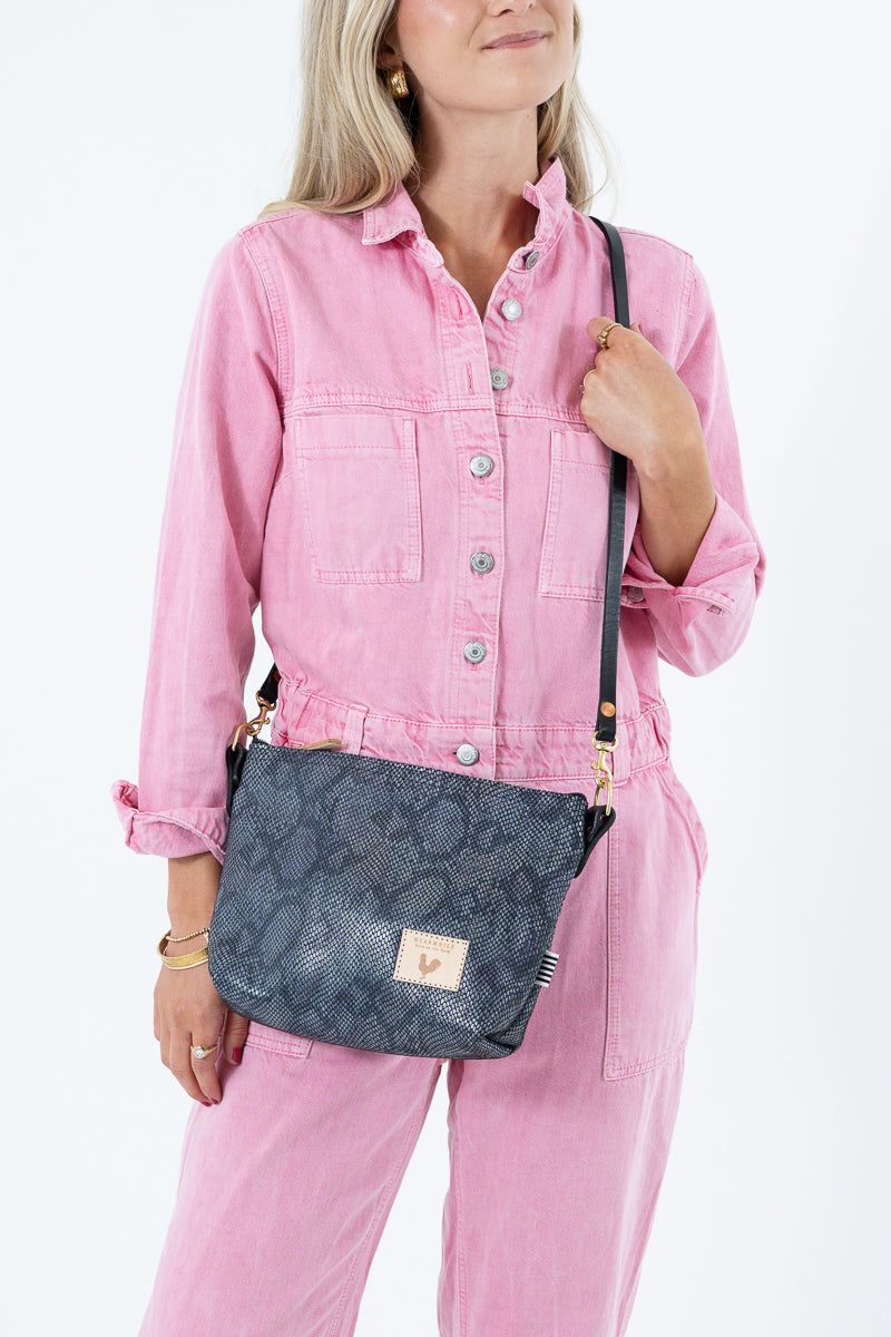 A woman wearing a pink jumpsuit and holding a dark gray snakeskin-patterned leather crossbody bag with a black strap and gold hardware, set against a plain white background. Handcrafted by Meanwhile Back on the Farm in Lynchburg, VA.