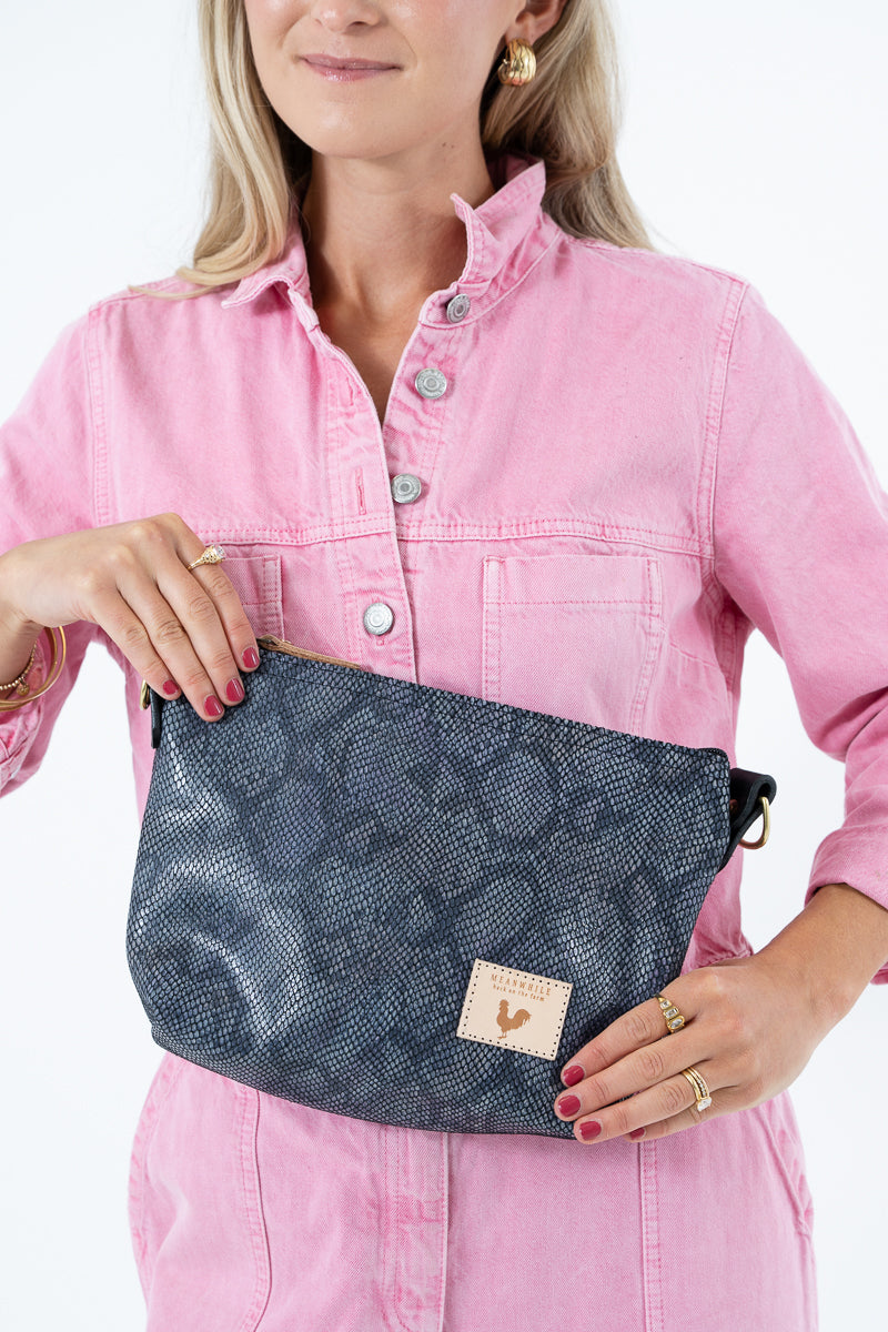 A woman wearing a pink jumpsuit and holding a dark gray snakeskin-patterned leather crossbody bag with a black strap and gold hardware, set against a plain white background. Handcrafted by Meanwhile Back on the Farm in Lynchburg, VA.