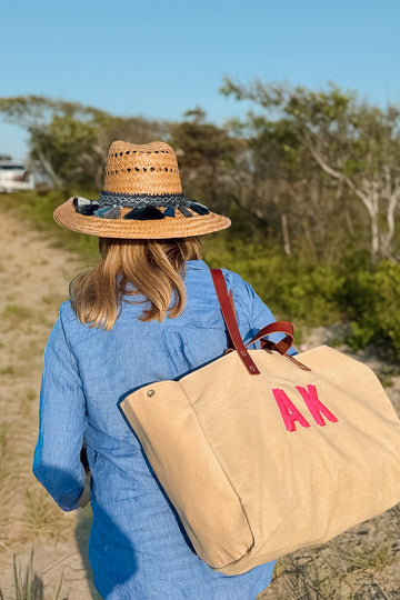 Woman at the beach holding a tan Meanwhile Back on the Farm tote bag with initials 'AK' in the center