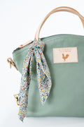A sage green pebbled leather handbag with a light tan top handle, a matching green shoulder strap, and a colorful floral print scarf tied in a bow around the handle. A beige leather patch on the front features the brand name. Handmade by Meanwhile Back on the Farm in Lynchburg,VA
