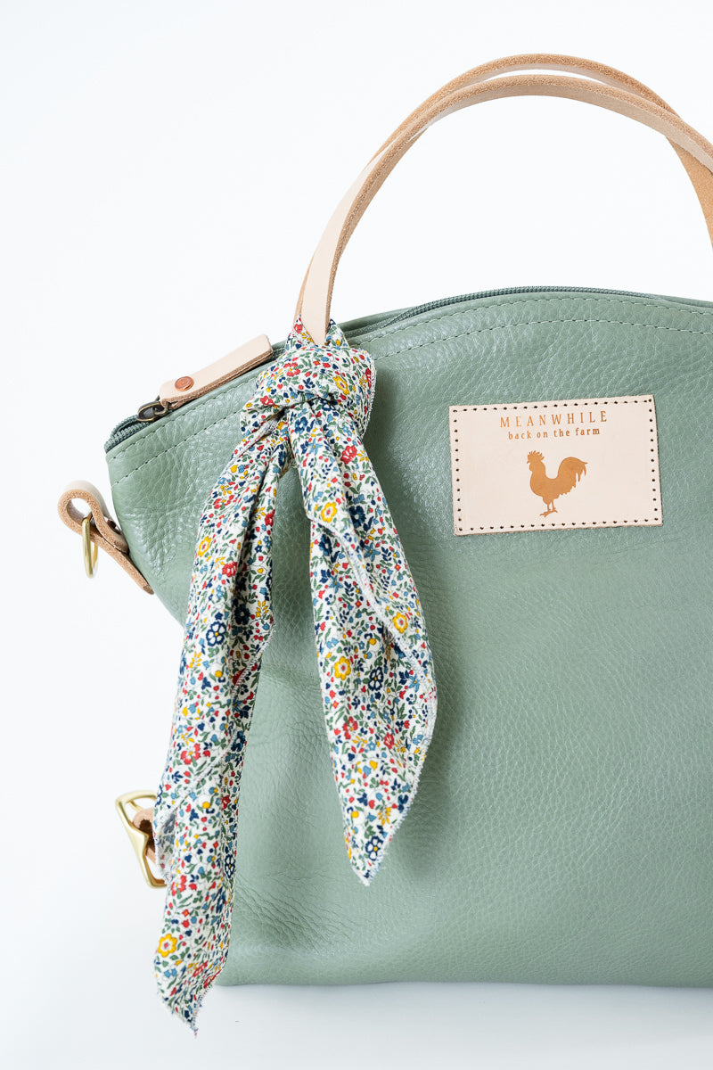 A sage green pebbled leather handbag with a light tan top handle, a matching green shoulder strap, and a colorful floral print scarf tied in a bow around the handle. A beige leather patch on the front features the brand name. Handmade by Meanwhile Back on the Farm in Lynchburg,VA