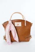 A brown leather tote bag with a light pink patterned scarf tied to its handle, set against a plain white background. The bag features a beige leather patch with a rooster logo. Handmade by Meanwhile Back on the Farm in Lynchburg,VA 