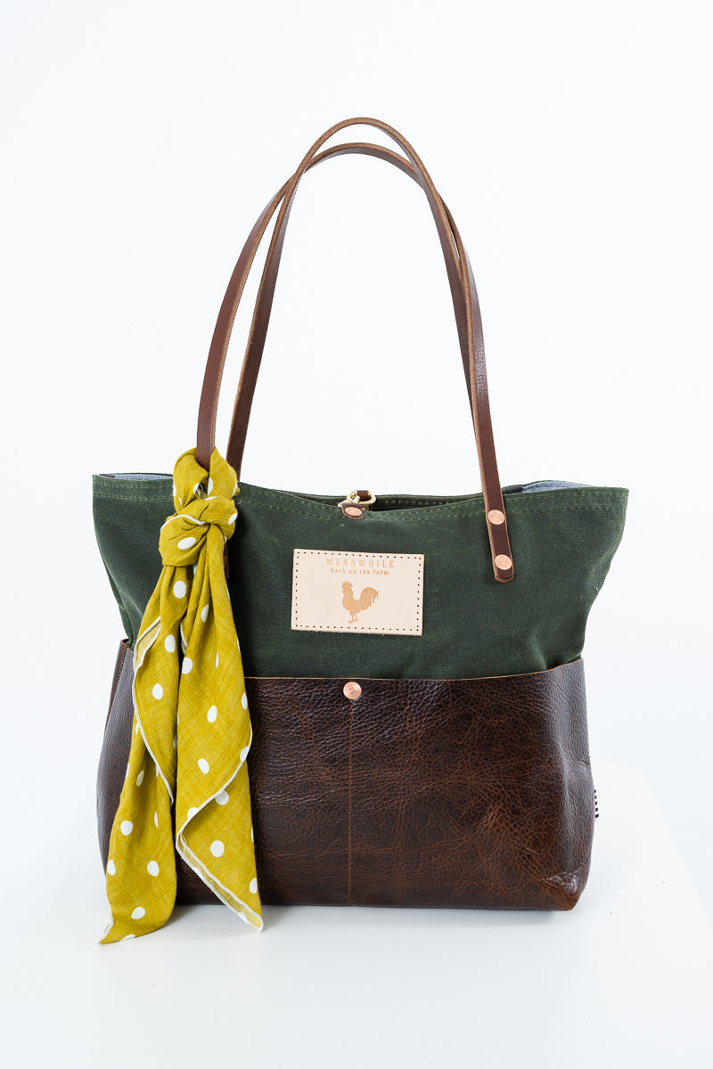 A two-tone tote bag with an olive green canvas top half and a dark brown leather bottom half, featuring brown leather shoulder straps and a small beige brand patch on the front. A saffron polka dot scarf is tied to the side of the bag. Handmade by Meanwhile Back on the Farm in Lynchburg,VA