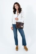 A woman modeling a dark brown leather shoulder bag with a tassel and a small gold logo plate, styled with a white long-sleeve blouse, blue jeans, and tan platform loafers against a white studio background. Handmade by Meanwhile Back on the Farm in Lynchburg,VA