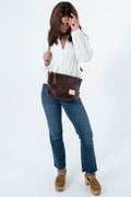 A woman modeling a brown leather crossbody bag with gold-tone hardware, tassel details, and a small rectangular brand patch, styled with a white blouse, blue jeans, and brown platform shoes against a white studio background. Handmade by Meanwhile Back on the Farm in Lynchburg,VA