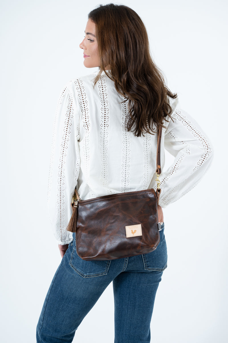 A woman wearing a white eyelet blouse and blue jeans is shown from the back and side, carrying a brown leather crossbody bag with gold-tone hardware and a small tan logo patch. Handmade by Meanwhile Back on the Farm in Lynchburg,VA