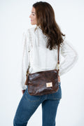 A woman wearing a white eyelet blouse and blue jeans is shown from the back and side, carrying a brown leather crossbody bag with gold-tone hardware and a small tan logo patch. Handmade by Meanwhile Back on the Farm in Lynchburg,VA