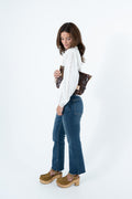 A woman posing in a white  long-sleeve top, blue flared jeans, and brown suede platform shoes, holding a brown leather shoulder bag against a plain white studio background. Handmade by Meanwhile Back on the Farm in Lynchburg,VA
