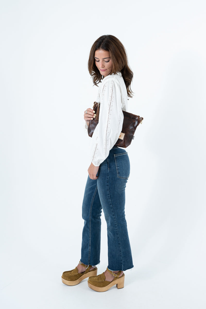 A woman posing in a white  long-sleeve top, blue flared jeans, and brown suede platform shoes, holding a brown leather shoulder bag against a plain white studio background. Handmade by Meanwhile Back on the Farm in Lynchburg,VA