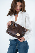 A woman wearing a white eyelet blouse and blue jeans holds a dark brown leather zip handbag with a tassel and a small tan brand patch. The background is a clean white studio backdrop. Handmade by Meanwhile Back on the Farm in Lynchburg,VA