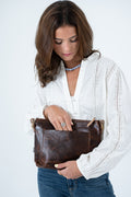 A woman wearing a white eyelet blouse and blue jeans holds a brown leather hand bag with gold-tone hardware against a plain white background. Handmade by Meanwhile Back on the Farm in Lynchburg,VA