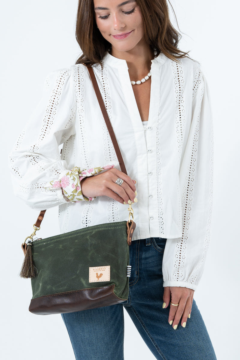 A woman wearing a white eyelet blouse and blue jeans carries an olive green waxed canvas and brown leather crossbody bag  a small leather patch on the front. Handmade by Meanwhile Back on the Farm in Lynchburg,VA