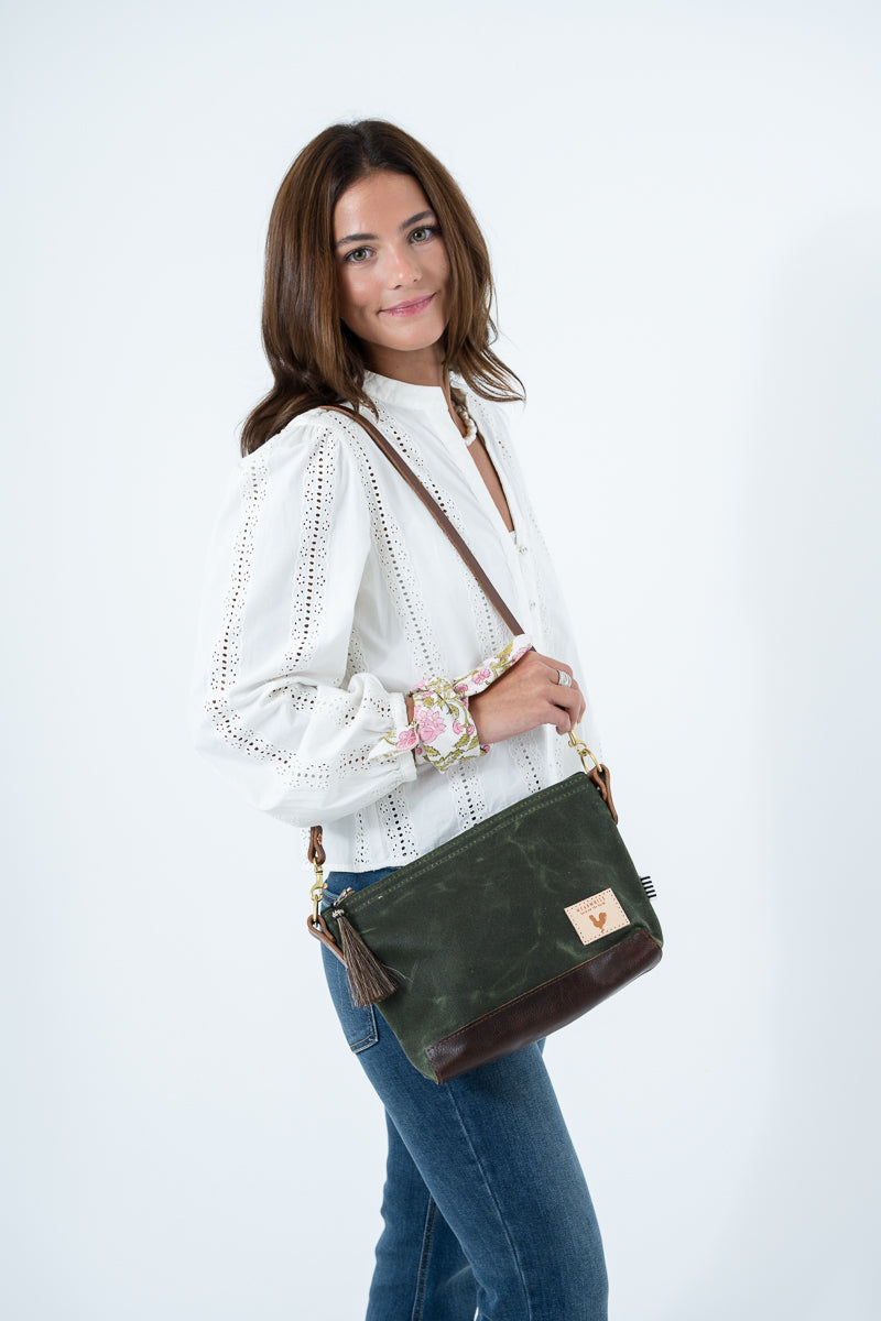 A woman wearing a white eyelet blouse and blue jeans carries an olive green waxed canvas and brown leather crossbody bag  a small leather patch on the front. Handmade by Meanwhile Back on the Farm in Lynchburg,VA