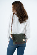 A woman shown from behind wearing a white eyelet blouse and blue jeans, carrying an olive green waxed canvas crossbody bag with brown leather trim and strap. Handmade by Meanwhile Back on the Farm in Lynchburg,VA