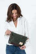 A woman wearing a white eyelet blouse and blue jeans holds a dark green leather handbag with a brown leather base and tassel detail against a plain white background. Handmade by Meanwhile Back on the Farm in Lynchburg,VA